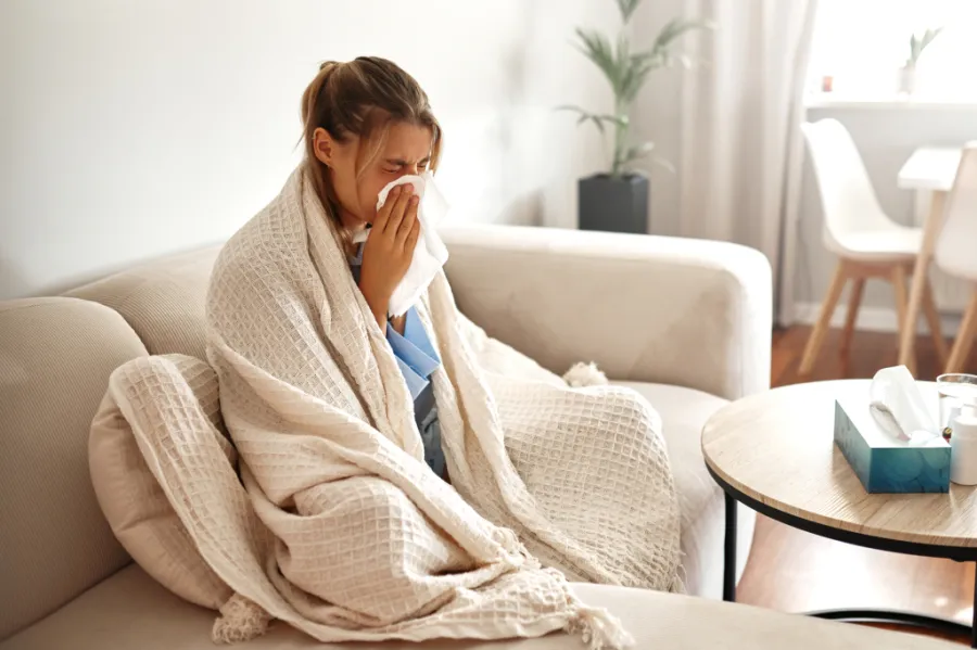 A sick person, wrapped in a blanket on the couch, blowing her nose