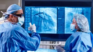 Cardiologist Omar Ali, MD, FACC in a heart and vascular surgery in Lowell General Hospital's Cath Lab talking to nurse while pointing to a large screen showing a heart scan.