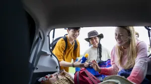 Friends wearing sun protection (long shirts, hats and sunscreen) are gathering their items from the back of their roadtrip car.