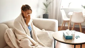 a sick person, wrapped in a blanket on the couch, blowing her nose