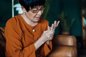 Senior woman rubbing her hands in discomfort from arthritis while sitting on sofa at home.