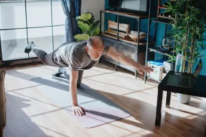 Senior joining in an online yoga class on his laptop while balancing on one foot and one hand on a yoga mat in their home.