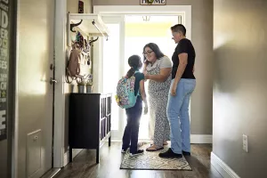 LGBTQ+ couple adjusting shirt collar of their child wearing a backpack as they stand in the front doorway of their home before the child goes off to school.