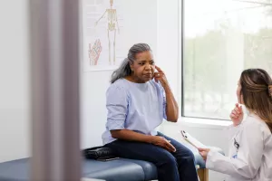Patient sitting on top of exam table while discussing ear symptoms with doctor during an audiology clinic appointment.