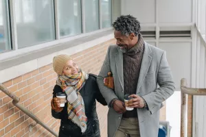 Senior couple bundled up in coats, scarves and hats and walking up the stairs to the train platform during a chilly autumn day holding to-go coffee cups and smiling at each other.