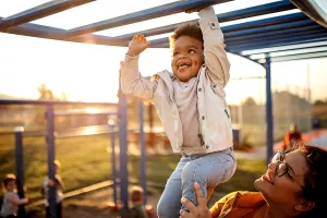 Child reaching and playing on the monkey bars in a playground in autumn with the help of their parent lifting them for support.