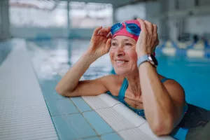 Senior wearing a swim cap and lifting up swimming goggles as they take a break from swimming in the community pool along the edge. 