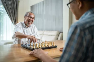 Seniors at home playing a game of chess at their dining room table and laughing.
