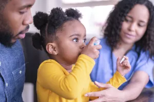 Nurse helping a toddler, pediatric patient, learn how to use an asthma inhaler during a clinic appointment while parent holds child.
