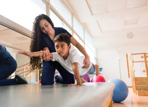 Physical therapist working with pediatric patient during clinic appointment and assisting child to lift arm and leg during a plank.
