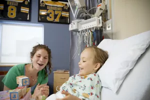 Pediatric patient in hospital bed at the Neely Pediatric Bone Marrow Transplant (BMT) unit at Tufts Medical Center with a Child-Life Specialist playing with wooden blocks.