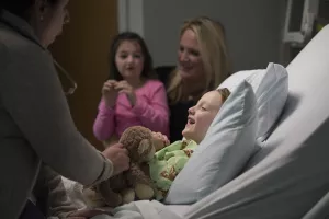 Nurse (Christine McEleney, RN) offers toy to inpatient child with mother and sister in the room at Tufts Medical Center.